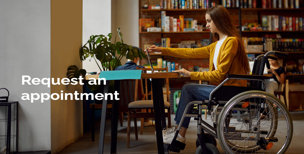 A woman in a wheelchair looking at a laptop on a table with a bookshelf full of books in the background. There is a sign 